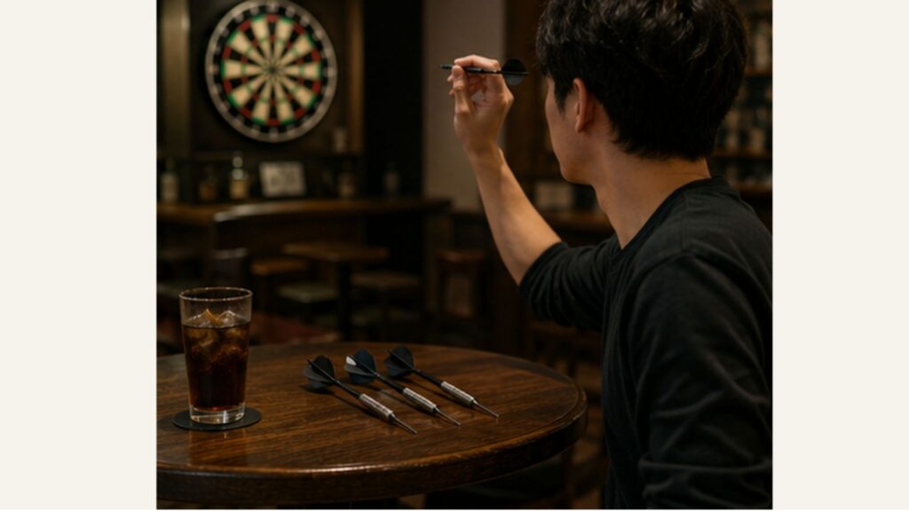 ダーツバーで手元のダーツを構え、テーブルにも予備のダーツを置いている男性